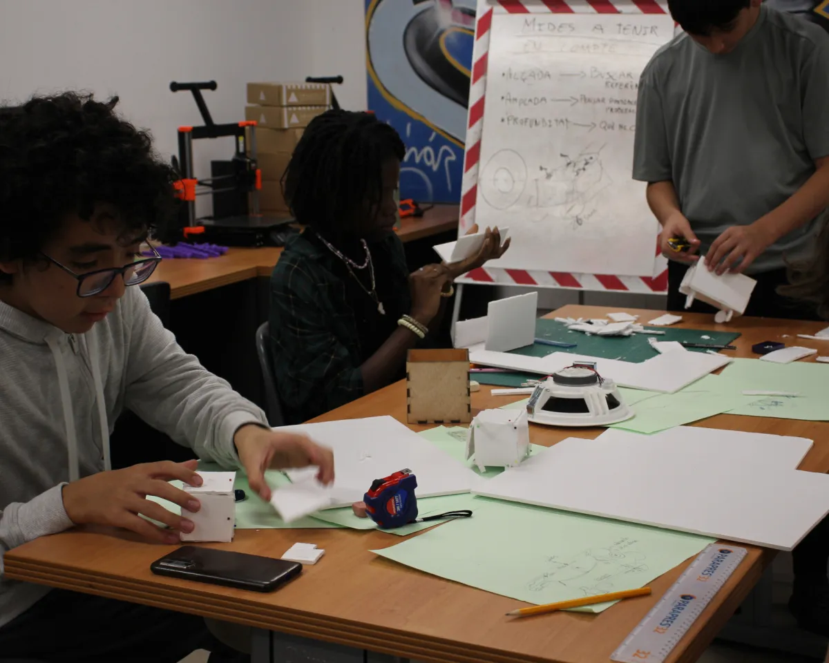 Un grup de joves treballa al voltant d'una taula. A sobre hi ha papers, mòbils, llapisos i una maqueta d'una capsa de fusta. 